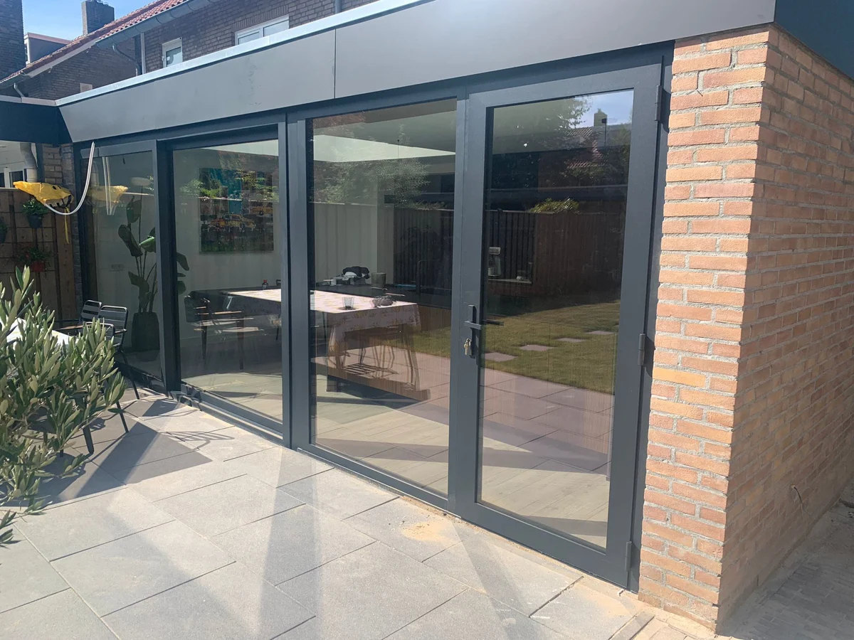 Aanbouw aan de tuin met brede glazen schuifdeuren, donkere kozijnen en een betegeld terras