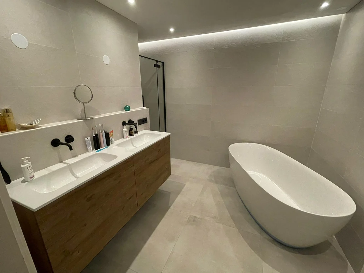 Badkamer met een wit vrijstaand bad, houten wastafelmeubel en indirecte plafondverlichting